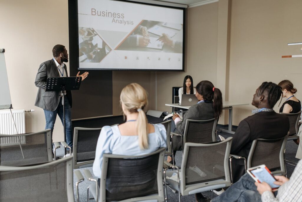 A diverse group attends a business analysis seminar with a presenter and projection screen in a modern conference room.